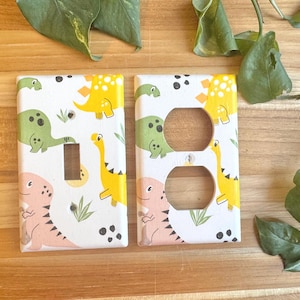 May include: Two white electrical outlet covers with a colourful dinosaur pattern. One cover has a toggle switch, the other has two outlet holes. The dinosaurs are yellow, green, and pink, with black spots and green leaves on a white background.