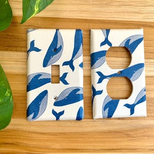 May include: Two white light switch and outlet covers with a blue whale pattern. The covers are made of plastic and have a matte finish.