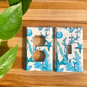 May include: Two decorative light switch covers with a coastal theme. The covers feature a white background with blue starfish, coral, and sand dollars. One cover has two outlet holes, and the other has a single switch opening.