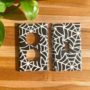 Könnte beinhalten: Zwei schwarze und weiße Lichtschalter- und Steckdosenabdeckungen mit einem Spinnennetz-Design.