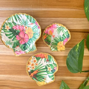 Op de afbeelding: Drie schelpvormige onderzetters met een tropisch bloemenontwerp. De onderzetters hebben roze, gele en oranje bloemen met groene bladeren op een witte achtergrond. De randen zijn goudkleurig afgewerkt. De onderzetters zijn gerangschikt op een houten oppervlak.
