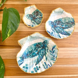 Peut inclure: Trois plats en céramique en forme de coquille de mer avec une bordure dorée, chacun représentant une peinture à l'aquarelle d'une tortue de mer sur un fond bleu et blanc.