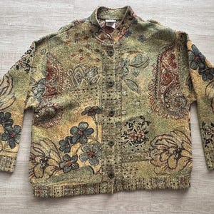 May include: A long-sleeve button-up jacket featuring a textured tapestry-style fabric with a multi-colored floral and paisley pattern in shades of olive green, gold, rust, and dark blue, finished with a stand-up collar and ribbed hem.