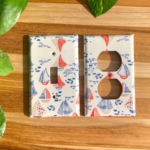 May include: Two white light switch covers with a nautical themed design. The design features red and blue sailboats on a white background.