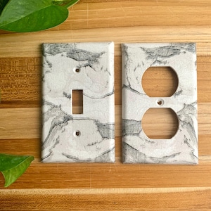Puede incluir: Dos cubiertas de enchufes eléctricos blancas con diseño de mármol. Una tiene una abertura para un interruptor, la otra tiene dos aberturas redondas para enchufes. Las cubiertas están sobre un fondo de madera.