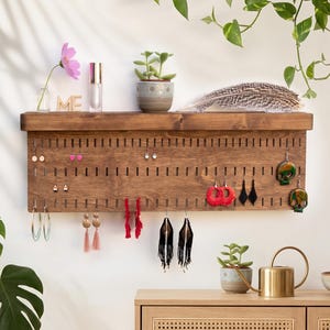 Puede incluir: Un organizador de joyas de pared rectangular de madera con filas horizontales de ranuras verticales para colgar pendientes. La unidad incluye un estante superior plano para exhibir pequeños objetos decorativos y accesorios.