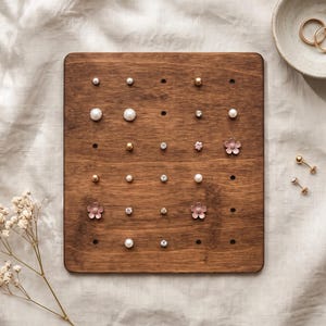 May include: A rectangular dark wood jewelry organizer board featuring a grid of holes populated with various stud earrings, including white pearls, gold spheres, clear crystals, and small pink flower-shaped studs, with two gold rings in a small bowl.
