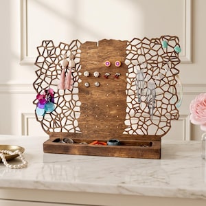 May include: A brown wooden jewellery organiser featuring a central perforated panel for earrings and two side panels with an intricate, organic web-like cutout pattern. The base includes a rectangular tray for small items and accessories.
