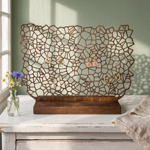 May include: A brown wooden jewelry display stand featuring an intricate, laser-cut geometric lattice pattern. The rectangular stand is mounted on a solid wooden base and holds several pairs of dangling earrings, including floral and pearl designs.