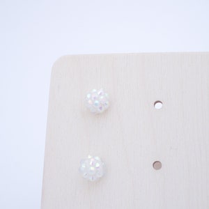 May include: A pair of iridescent white stud earrings displayed on a wooden earring holder.