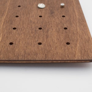 May include: A dark brown wooden board with evenly spaced holes for holding jewelry. The board has a smooth, polished finish.