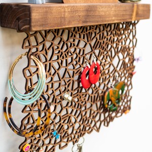 May include: A wooden wall-mounted jewelry organizer with a geometric design. The organizer features a shelf at the top and a decorative lattice pattern with multiple hooks for hanging earrings, necklaces, and other jewelry.