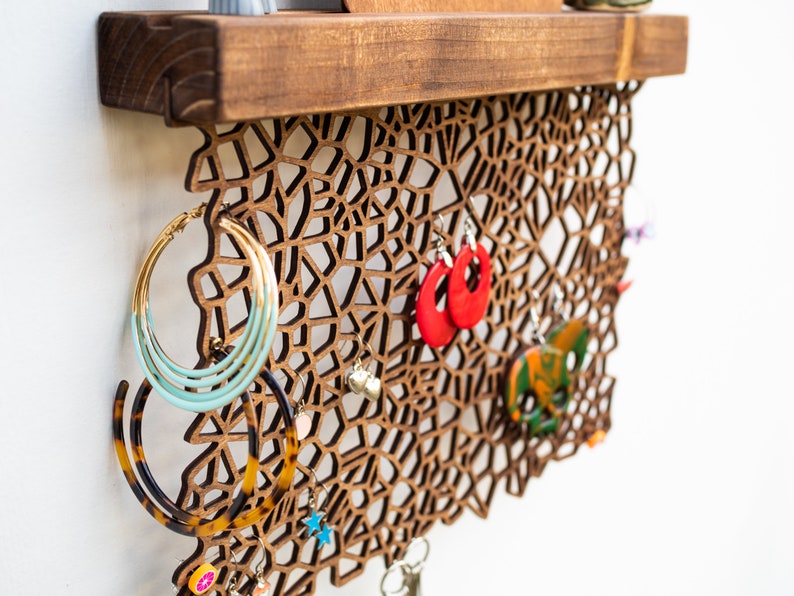 May include: A wooden wall-mounted jewelry organizer with a geometric design. The organizer features a shelf at the top and a decorative lattice pattern with multiple hooks for hanging earrings, necklaces, and other jewelry.