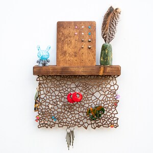 May include: A wooden shelf with a geometric design, featuring a small blue toy, a green vase with a feather, and a corkboard with colorful push pins. The shelf is decorated with various earrings, keys, and a small elephant figurine.