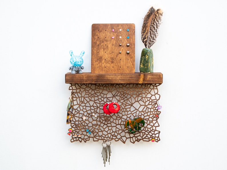 May include: A wooden shelf with a geometric design, featuring a small blue toy, a green vase with a feather, and a corkboard with colorful push pins. The shelf is decorated with various earrings, keys, and a small elephant figurine.