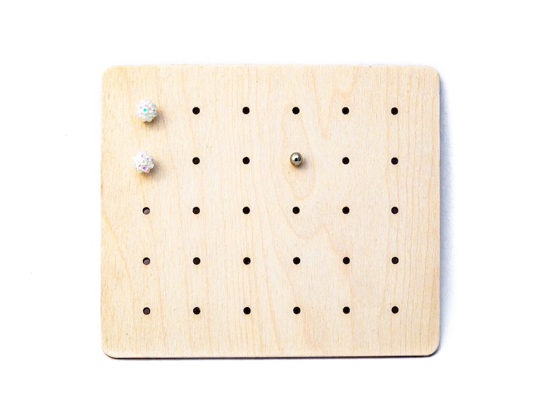 May include: A wooden earring display board with holes for holding earrings. There are two white sparkly earrings and a silver bead on the board.