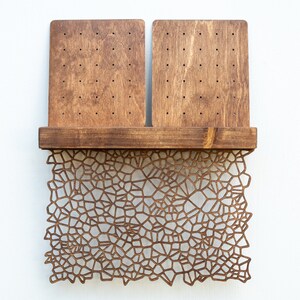 May include: A wooden earring holder with two rectangular panels and a shelf. The panels have multiple holes for holding earrings. The shelf is decorated with a laser-cut design of a geometric pattern.
