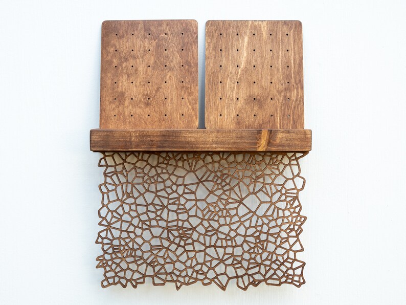 May include: A wooden earring holder with two rectangular panels and a shelf. The panels have multiple holes for holding earrings. The shelf is decorated with a laser-cut design of a geometric pattern.