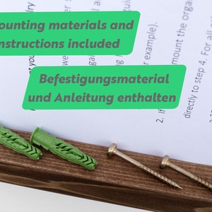 May include: Two green plastic wall anchors and two silver screws on a brown wooden surface. The text "Mounting materials and instructions included" and "Befestigungsmaterial und Anleitung enthalten" is visible on a white sheet of paper.