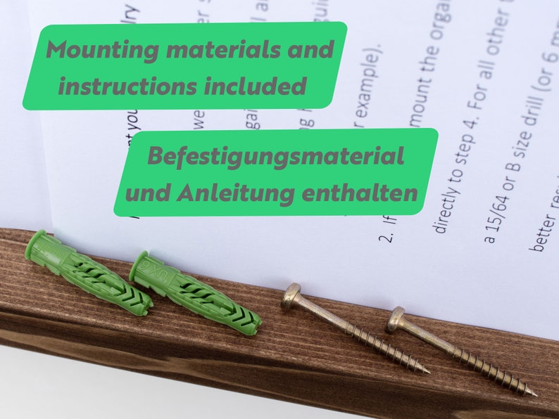May include: Two green plastic wall anchors and two silver screws on a brown wooden surface. The text "Mounting materials and instructions included" and "Befestigungsmaterial und Anleitung enthalten" is visible on a white sheet of paper.