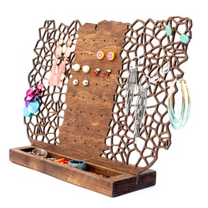 May include: Wooden jewelry stand with a geometric design. The stand has multiple holes for earrings and a small shelf for rings. The stand is brown and has a natural wood finish.