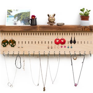 Puede incluir: Un organizador de joyas de madera con un estante en la parte superior. El organizador tiene una placa perforada con agujeros para pendientes y ganchos para collares. Hay varios pendientes y collares colgados del organizador.