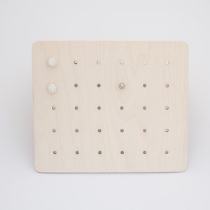 May include: A wooden earring display stand with multiple holes for storing earrings. The stand is light brown and has two white pearl earrings and one silver earring.