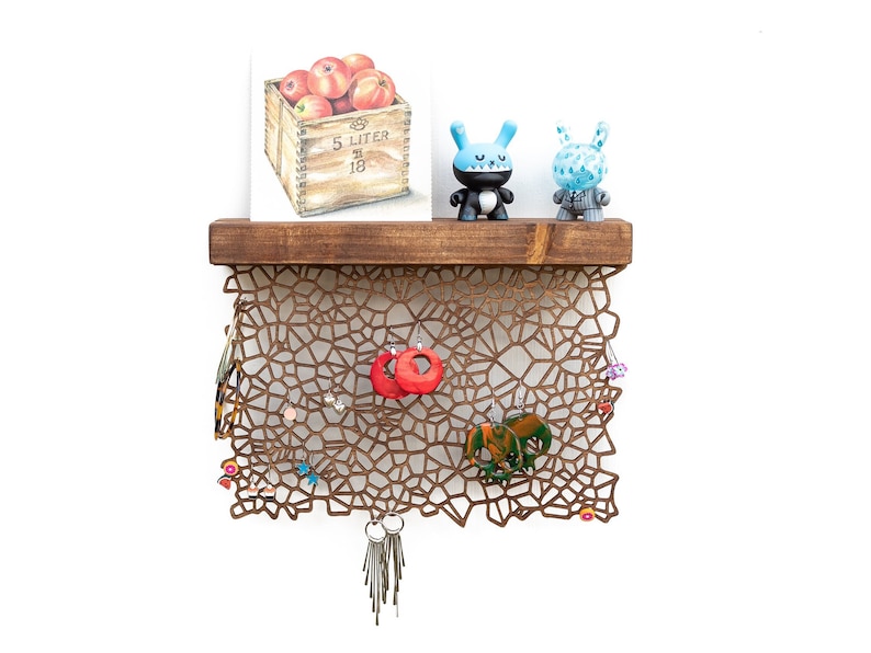 May include: A wooden shelf with a geometric metal design hanging below. The shelf has a painting of apples in a crate on it and two blue toy figures. The metal design has various earrings hanging from it.