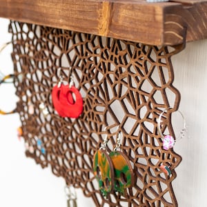 May include: Wooden earring holder with a geometric design. The holder has a shelf at the top and is decorated with various earrings in different colors and styles.