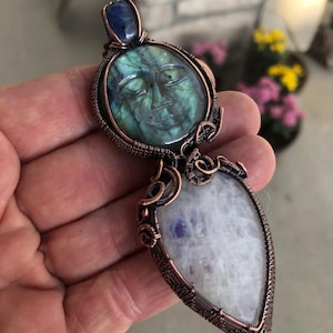 Peut inclure: Pendentif artisanal avec un visage en labradorite sculptée et un fil de cuivre. Le pendentif comprend une pierre de lune en forme de goutte et un accent de pierre bleue. Le pendentif mesure environ 7.6 cm de long.