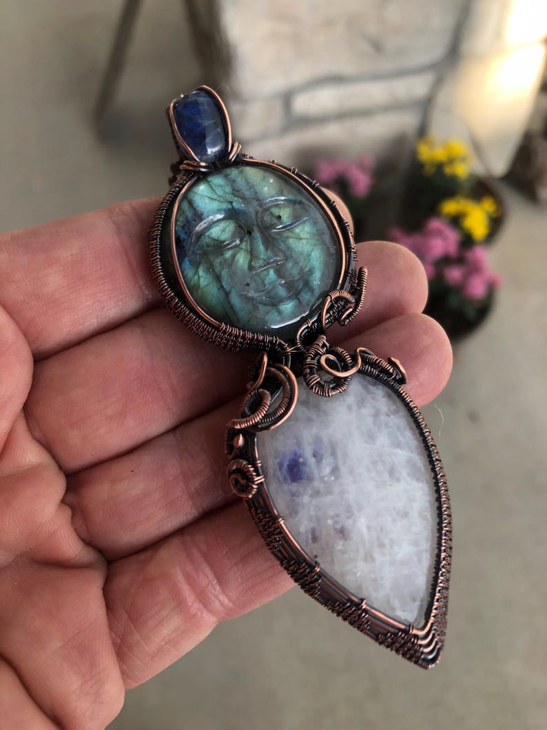 Op de afbeelding: Handgemaakte hanger met een gebeeldhouwd labradoriet gezicht en een koperdraad wikkel. De hanger bevat een druppelvormige maansteen en een blauwe edelsteen accent. De hanger is ongeveer 7.6 cm lang.