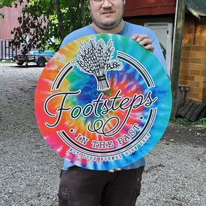 May include: A person holding a round, tie-dye sign with the text "Footsteps in the Past. Not your normal sign shop." The sign has a rainbow color scheme with a black wheat sheaf design.