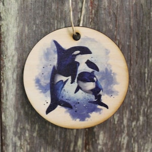 May include: A round wooden ornament with a watercolor-style image of two orcas swimming in a blue and white background.