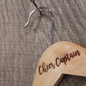 May include: A wooden clothes hanger with a metal hook. The hanger is engraved with the words "Cheer Captain".
