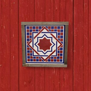 May include: Square framed artwork with a geometric star design in red, white, and blue. The artwork is set against a red wooden background. The frame is a natural wood color.