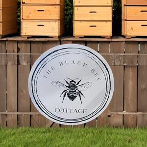 May include: Wooden beehives with multiple stacked boxes sit behind a wooden fence. A round sign in the foreground reads "THE BLACK BEE COTTAGE" with a bee illustration in the center. The sign is white with black text and a black border.