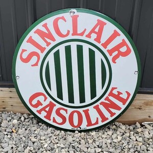 May include: A round Sinclair Gasoline sign with a white background, green border, and red lettering. The center features green and white vertical stripes. The sign has holes for mounting and is displayed outdoors.
