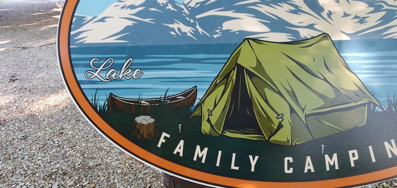 May include: A colorful oval sign with a mountain scene, a lake, a canoe, a green tent, and the text "Lake Family Camping".