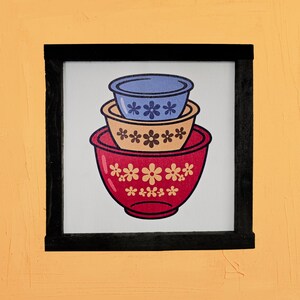 May include: A framed print featuring a stack of three mixing bowls. The top bowl is blue, the middle is tan, and the bottom is red. Each bowl is decorated with white flower patterns. The frame is black, and the background is a light orange.