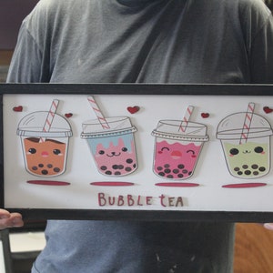 May include: A framed wooden sign with a white background and four cartoon bubble tea cups with different colored drinks and smiling faces. The sign says "BUBBLE TEA" in red letters.