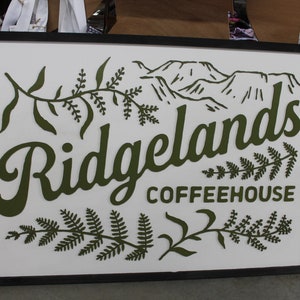 May include: A framed sign with a white background and black frame. The sign features a stylized green fern design and the text "Ridgelands Coffeehouse" in green.