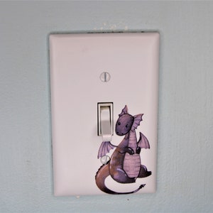 May include: A white light switch cover with a purple and gray dragon illustration. The dragon is sitting on its haunches with its wings spread out.