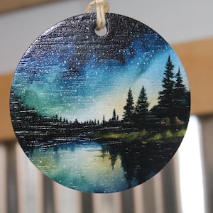 May include: A round wooden ornament with a painted scene of a starry night sky over a lake and forest. The sky is a deep blue with green and purple hues, and the stars are white. The lake is a dark blue with a reflection of the sky. The forest is a dark green.