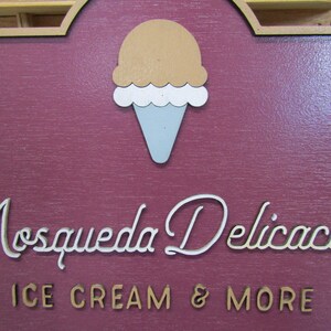 May include: A wooden sign with a brown background and gold trim. The sign has a white ice cream cone with a brown scoop of ice cream on top. The text "Mosqueda Delicacies" is written in white cursive lettering with a gold outline. Below the text is "ICE CREAM & MORE" in gold lettering.