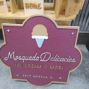 May include: A maroon wooden sign with a gold border and a white cursive font reads "Mosqueda Delicacies Ice Cream & More" with a small ice cream cone graphic above the text. The sign also includes the text "Fort Morgan, CO" in a smaller gold font.