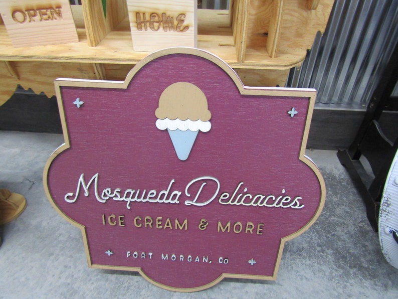 May include: A maroon wooden sign with a gold border and a white cursive font reads "Mosqueda Delicacies Ice Cream & More" with a small ice cream cone graphic above the text. The sign also includes the text "Fort Morgan, CO" in a smaller gold font.