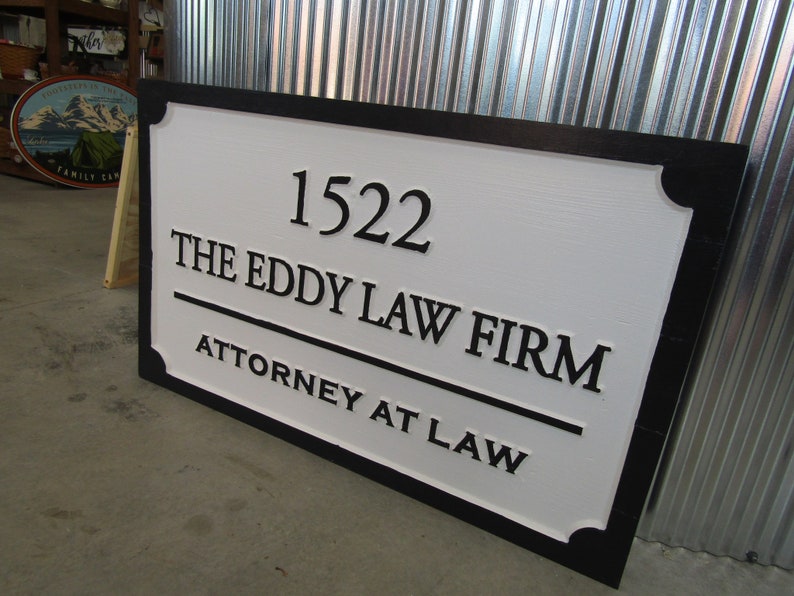 Puede incluir: Un letrero de madera blanco con borde negro y letras en relieve. El letrero dice "1522 The Eddy Law Firm Attorney at Law".