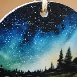 May include: Round wooden ornament with a painted image of a starry night sky and a silhouette of trees in the foreground.