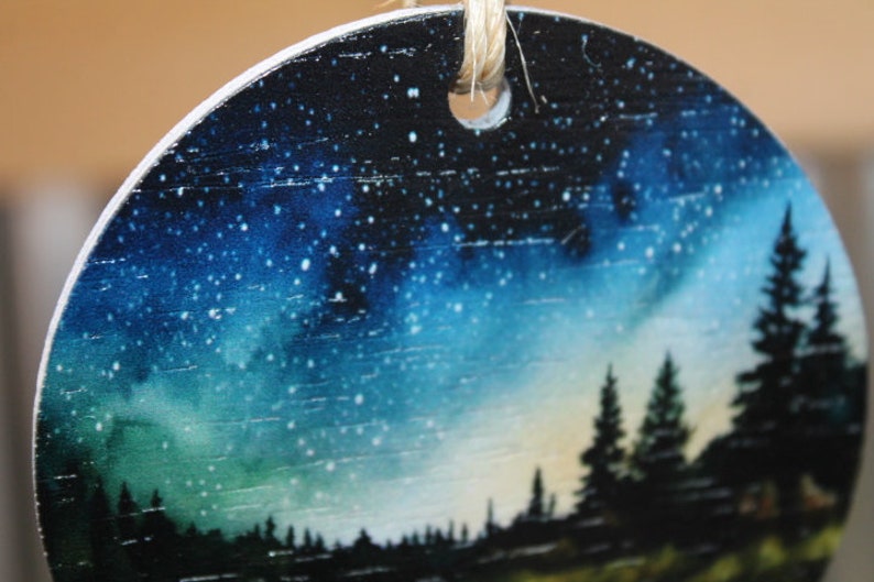 May include: Round wooden ornament with a painted image of a starry night sky and a silhouette of trees in the foreground.