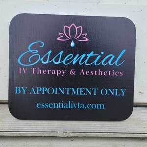 May include: Black rectangular sign with rounded corners. The sign features the words "Essential IV Therapy & Aesthetics" in blue and pink, with a lotus flower graphic. The text "BY APPOINTMENT ONLY" and a website address are also displayed.